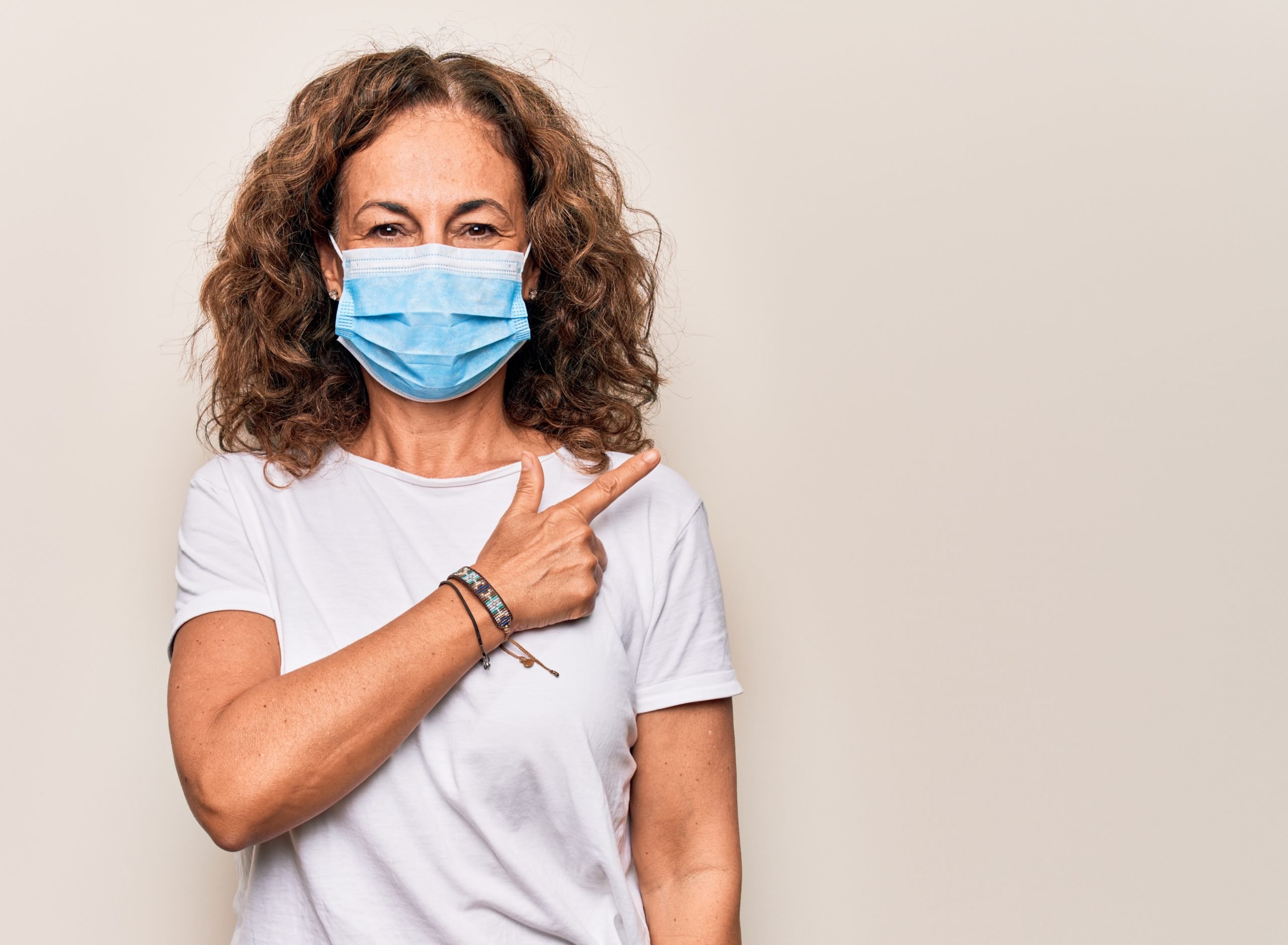 A woman with curly brown hair wearing a blue mask on a beige background.