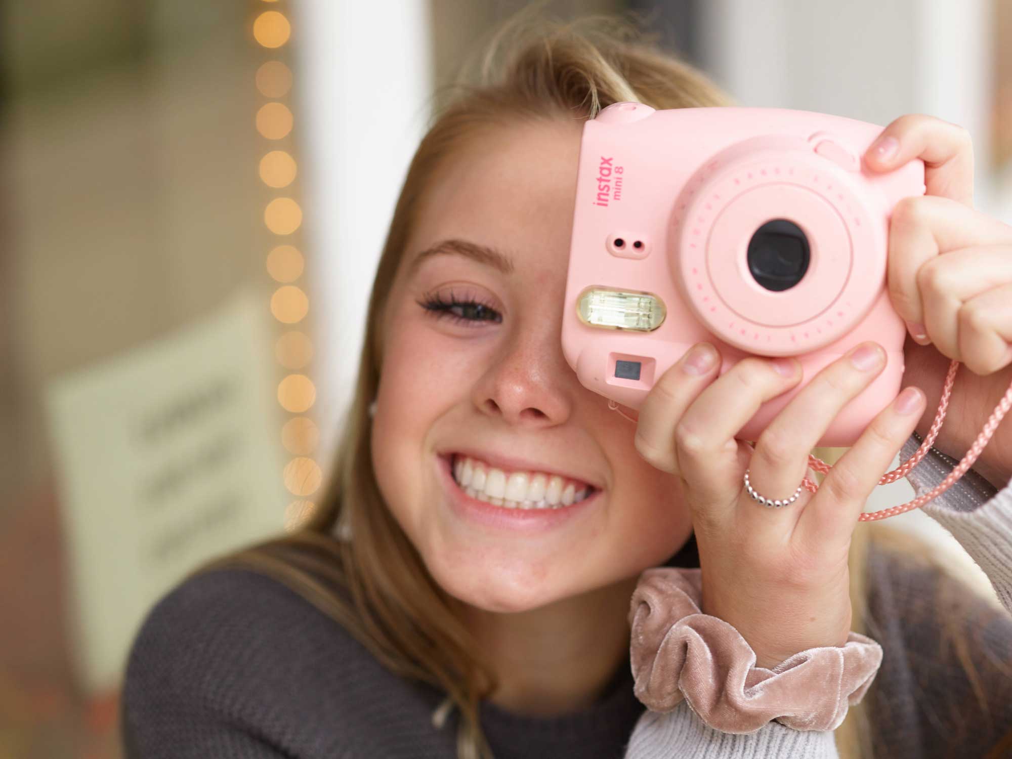 A teen girl with red hair smiling and taking a photo with a Polaroid camera.